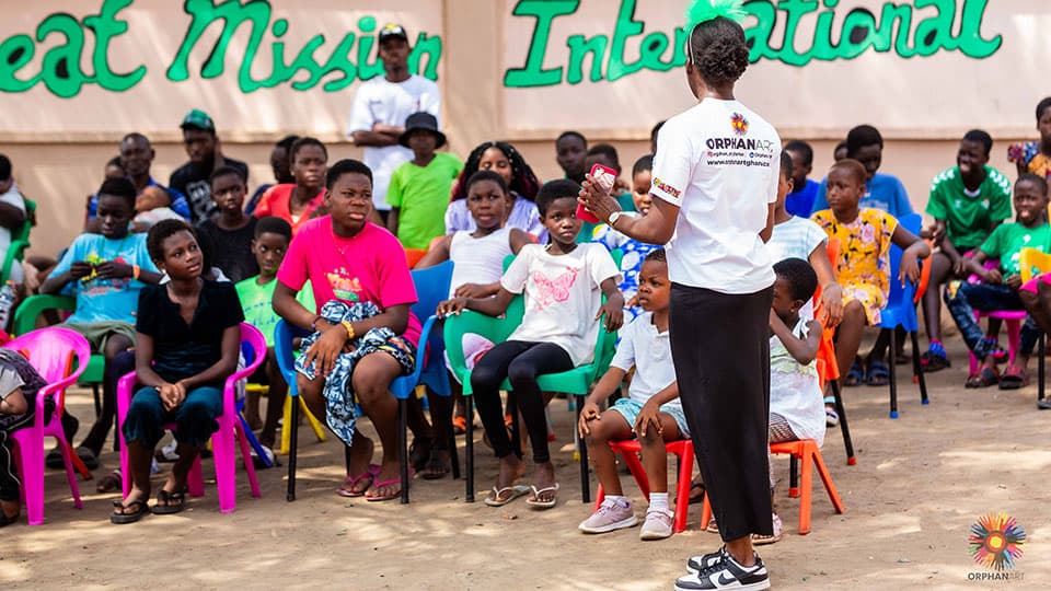 A group of children and young adults in Ghana. They are all pointing towards and listening to someone who is speaking in a white t-shirt that reads Orphan Art.